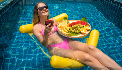 Beautiful woman lying on floating hammock in the swimming pool with wine and tray of fruits