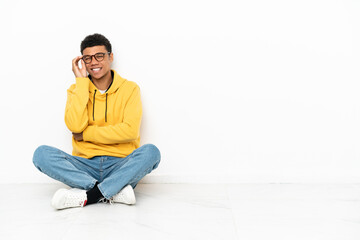 Young African American man sitting on the floor isolated on white background with glasses and happy