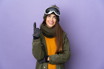 Wall Mural - Skier caucasian girl with snowboarding glasses isolated on purple background pointing up a great idea