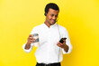 © luismolinero - African American Telemarketer man working with a headset over isolated yellow background holding coffee to take away and a mobile