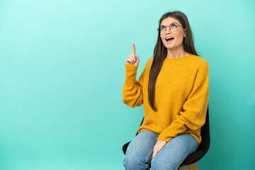 Wall Mural - Young caucasian woman sitting on a chair isolated on blue background pointing up and surprised