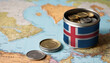 A jar filled with cash, displaying the Iceland flag, sits atop a map. Saving for vacation, leisure activities. Financial planning, travel budget allocation