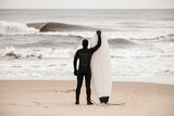 Fototapeta  - Slender male surfer stands on the seashore and holds his surfboard