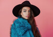 Full body photo of an beautiful woman, wearing black top and hat with long dark brown hair in big fluffy blue fur coat posing on pink background