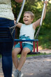 A happy child is sitting on a swing and is ready to swing high, he is looking at his mother with joyful and impatiently eyes