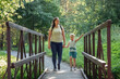 Mother and son hand in hand walking together on wooden bridge in sunny summer forest. Horizontal photo