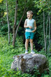 A boy stands on a large stone in green summer forest nature background.