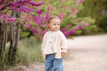 Wall Mural - Stylish baby girl 1-2 year old wearing trendy casual clothes over flowers on blooming tree in park outdoor. Summer season.