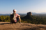 Fototapeta Sawanna - Man with backpack sitting on rock and looking at landscape. Beautiful scenery with Sigiriya rock. Solo traveler in Sri Lanka. .