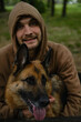 A young guy is sitting in a spring forest and hugging a German shepherd. Male owner with a dog on a walk in a green park. Front view portrait of two best friends.