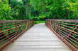 A rustic wooden bridge, adorned with rust-colored railings, beckons entry into a wooded forest sanctuary adorned with thick foliage.