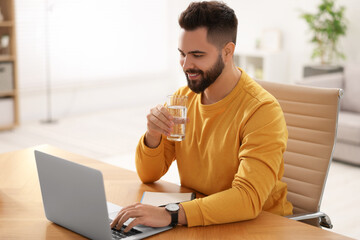 Poster - Young man with glass of water watching webinar at table in room