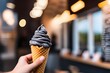 A hand holding a chocolate ice cream cone with a charcoal soft serve, against a blurred background.