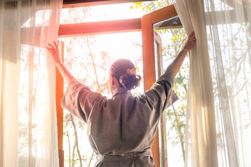 Wall Mural - The woman was at the window in the bedroom. She opened the curtains on the window. In the morning and she looks at the view of mountains and trees at sunrise.	