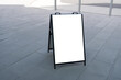 A mockup template of a blank white easel board stands on an urban walkway. An empty background texture of an A-Frame sandwich sign with copy space on the pedestrian sidewalk. 