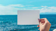 © Ruslan - A woman's hand holds a white blank piece of paper against the background of the sea. Mockup, product advertisement, invitation flyer