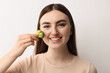 Smiling woman making fake freckles with broccoli and cosmetic product on light background