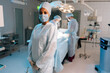 Portrait of skilled African-American female doctor standing posing looking at camera in light operating theater. Diverse team of surgeons performing operation on background. Concept of emergency.