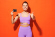 Young fitness trainer instructor sporty woman sportsman wear purple top spend time in home gym hold credit bank card show thumb up isolated on plain orange background. Workout sport fit abs concept.