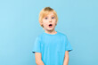 Wow! Portrait of a shocked cute little boy with blond hair covering open mouth with one hand, on pastel blue background. Surprised preschooler studio shot.