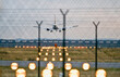 Landung eines Passagierflugzeugs auf einem Flughafen mit den erleuchteten, unscharf fotografierten Positionslampen der Landebahn und Stacheldrahtzaun im Vordergrund