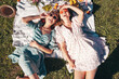 Two young beautiful smiling hipster female in summer sundress and hats. Carefree women making picnic outside. Positive models sitting on plaid on grass, drinking champagne,  eating fruits. Top view