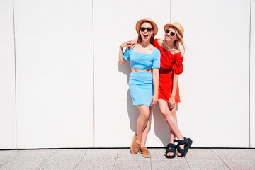 Two young beautiful smiling hipster women in trendy summer same clothes. Carefree models posing in the street. Positive models having fun. Cheerful and happy. In hat and sunglasses. Near wall