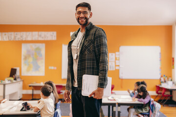 Wall Mural - Happy elementary school teacher standing in a classroom, with children in the background