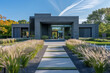 A modernist residence with a slate gray exterior, surrounded by an artistic arrangement of native plants and grass. It features a glass-enclosed atrium, solar shades, and an automated entry system