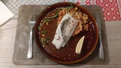 On a wooden table with a tablecloth is a leather lined table and on it stands a ceramic plate with a baked fish fillet and rice porridge with vegetables. A metal fork and knife lie nearby. Dinner time