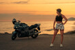 Caucasian young woman in daring clothes poses near a tourist motorcycle. Golden sunset in the background. The concept of the World Motorcyclist Day