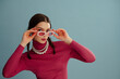 Fashionable confident woman wearing trendy rectangular pink sunglasses, turtleneck, pearl necklace, posing on blue background. Studio fashion portrait. Copy, empty, blank space for text