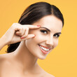 Skin care beauty concept. Portrait of happy smiling woman pointing showing brow or eye, touching skin, isolated against orange yellow background. Brunette model at studio. Square image.