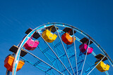 Fototapeta Las - Colorful ferris wheel on a fairground in California. Orange, yellow and pink gondola cabins isolated on blue sky. Historic amusement park “Boardwalk“ on the beach of Santa Cruz, California (USA)