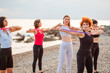 Group of Caucasian adult women in sportswear are training with sports elastic band at beach. Sea is in background. Concept of sport and fitness