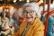 © Helena GARCIA - Mujer senior de 70 años con gafas graduadas, sentada sonriente en una atracción de feria, sobre fondo desenfocado de carrusel o tiovivo de caballitos