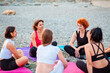 Female yoga class. Group of happy young-adult multi-racial women are sitting on sports mats on wild beach and talking to each other. Concept of female circle of communication and support