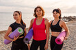 Portrait of group of Caucasian pretty adult women holds sports mats on pebble beach. Sea and wild coast in background. Fitness outdoor training