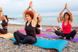 Outdoor activity at sea coast. Side view of group of Caucasian women meditate sitting on sports mats on pebble beach. Yoga asana for meditating