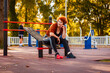 Mid adult Caucasian smiling woman is sitting on playground and listening to music with headphones. Autumn park in background. Copy space. Concept of fitness and training