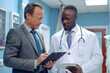 © GustavsMD - An African doctor in a white lab coat and a businessman in a suit analyze data together on a tablet in a modern hospital setting, symbolizing collaboration in healthcare