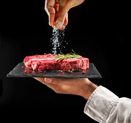 Wall Mural - Chef is salting or seasoning raw ribeye steak laying on graphite serving board. Black background.