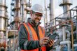 © Monktwins - Smiling middle-aged male engineer in protective helmet and safety vest holds tablet computer. Oil production plant at background.