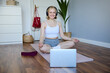 Portrait of fit and healthy woman at home, practice yoga, sitting on rubber mat, listening to instructions online, using meditation music to relax, following guidance on laptop