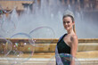 Portrait of young, pretty, blonde woman in green sequined party outfit, with diamond crown and beauty pageant winner's sash, posing next to a fountain surrounded by soap bubbles.