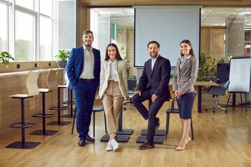 Wall Mural - Happy corporate business team in office. Group portrait of happy, smiling, cheerful people in classic suits standing by table in conference room with projection screen in background