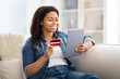 © Prostock-studio - Woman Sitting on Couch Holding Credit Card and Tablet