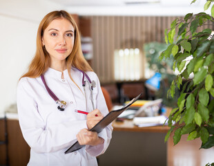 Wall Mural - Young woman therapist working in a medical clinic writes important notes on a tablet, standing in a well-lit office. Close-up ..portrait