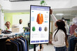 Female customer reviewing clothes to purchase from self ordering kiosk service, looking at trendy fashion items on touch screen board. Young client checking products on monitor.