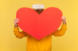 Portrait of angelic anonymous woman with nimb above head with Afro hairstyle hiding behind big red heart, wearing casual style hoodie. Indoor studio shot isolated on yellow background.